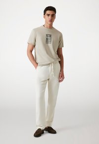Beige katoenen t-shirt met een grafische print, gecombineerd met crèmekleurige broek met trekkoord. Bruine instapschoenen maken de casual outfit compleet.