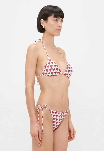 Traiņķa bikini baltā krāsā ar sarkanu sirds rakstu; satur regulējamus saitītes un aukliņas, uzsverot vienkāršu dizainu un kopējo jautrību.