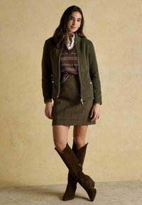 Žena oblečená v olivovozelenej bunde cez vzorovaný sveter, károvanú sukňu a hnedé semišové topánky po kolená, stojaca pri béžovej stene.