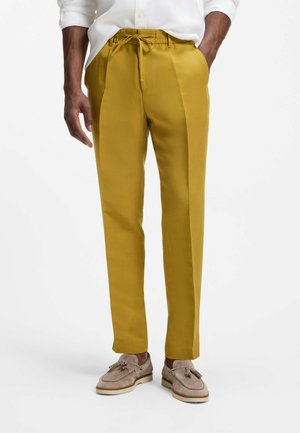 Uomo che indossa pantaloni giallo senape su misura con coulisse in vita, camicia bianca e mocassini beige con nappine, mani in tasca.
