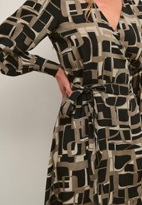 Schwarzes und beiges Wickelkleid mit geometrischem Muster, langen Ärmeln, V-Ausschnitt und Taillengürtel für eine figurbetonte Silhouette. Glatte Textur.