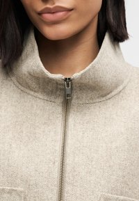 Beżowa kurtka z szewronowym wzorem zapinana na zamek, z wysokim kołnierzem; posiada metaliczny zamek i miękką fakturę tkaniny. Brak widocznych wzorów ani akcentów.