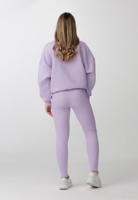 Fialová mikina se spadlými rameny spojená s odpovídajícími ribkovanými legínami. Bílé tenisky doplňují outfit. Hladká textura látky.