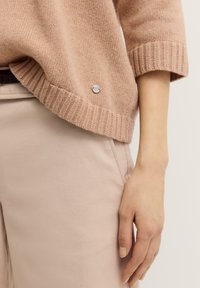 Helles rosa gestricktes Sweatshirt mit einem gerippten Saum, versehen mit einem kleinen Logoakzent an der Seite. Kombiniert mit beigen maßgeschneiderten Hosen.