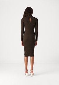 Robe marron à manches longues et côtelée, longueur mi-mollet, avec une découpe en forme de clé à l'arrière ; associée à des sandales à talons blocs blanches.