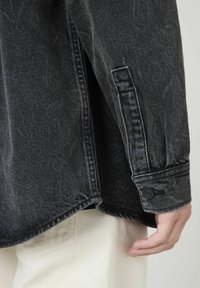 Veste en jean noire avec une finition texturée, des coutures visibles et un poignet à boutons. Portée sur un pantalon de couleur claire, mettant en valeur des styles superposés.