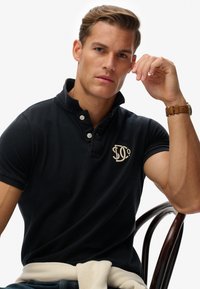 Marineblaues Polohemd mit Kragen und zwei Knöpfen. Mit einem gestickten Logo auf der Brust. Modell sitzt mit einem leichten Pullover auf dem Schoß.