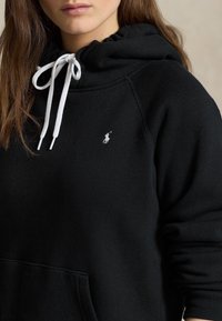 Černá mikina s kapucí a bílou šňůrkou, s malým vyšitým logem na hrudi. Má měkkou tkaninu a klokaní kapsu.