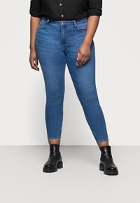 Persona vistiendo jeans ajustados azules de tiro medio, botas negras hasta el tobillo y una camisa negra de botones con las mangas arremangadas.