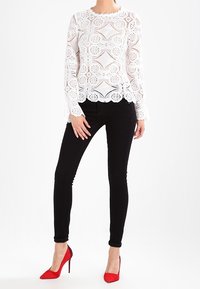 Valkoinen pitsipaita geometristen kuvioiden ja aallotetuilla reunoilla, yhdistettynä mustiin kapeisiin farkkuihin ja punaisiin korkokenkiin.