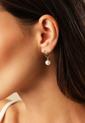 Goldene Creolen mit unregelmäßiger ovaler Form, die eine hängende Perle aufweisen, vor einem neutralen Hintergrund präsentiert.