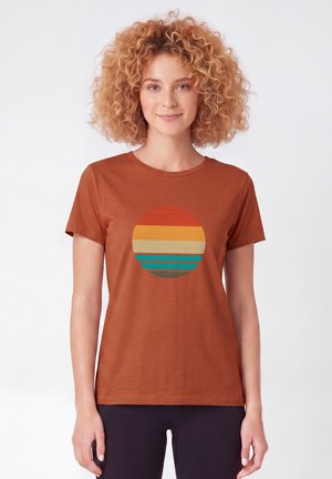 Femme aux cheveux bouclés portant un t-shirt couleur rouille avec un cercle de rayures horizontales orange, jaune, beige, sarcelle et marron.