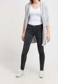 Světlě šedý pletený kardigan s kapucí přes bílý tílko, doplněný černými úzkými džíny a bílými teniskami. Celkově minimalistický design.