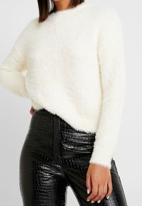 Weißer, flauschiger Pullover mit rundem Ausschnitt, langen Ärmeln und gerippten Bündchen, kombiniert mit schwarzen, glänzenden, kunstledernen Hosen im Krokodil-Look.