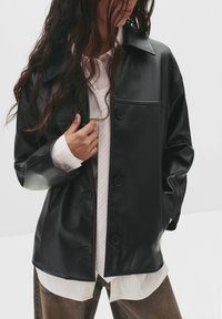 Schwarze Kunstlederjacke mit klassischem Kragen, Knopfleiste und übergroßem Schnitt, über einem gestreiften weißen Hemd getragen.