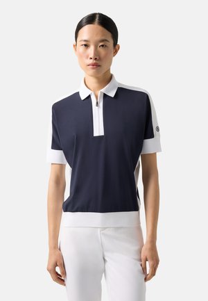 Frau mit schwarzem Haar in einem glatten Pferdeschwanz, die ein marineblaues Poloshirt mit weißem Kragen und weißen Ärmeln trägt, kombiniert mit einer weißen Hose.