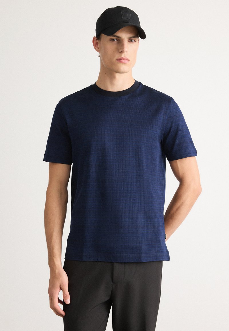 Homem vestido com uma camisa de mangas curtas texturizada azul-marinho, calças pretas e um boné de baseball preto, de pé contra um fundo branco.