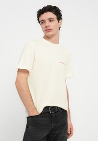 Κρεμ βαμβακερό t-shirt με κοντά μανίκια, στρογγυλή λαιμόκοψη και κόκκινο κεντημένο λογότυπο στο στήθος. Φορεμένο με μαύρο τζιν τζιν και μαύρη ζώνη.