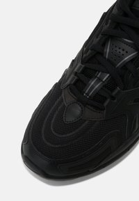 Černá sportovní bota s vrchním dílem z prodyšné síťoviny, gumovými doplňky, texturovanými vzory a kontrastními lesklými detaily podél stran a šňůrek.