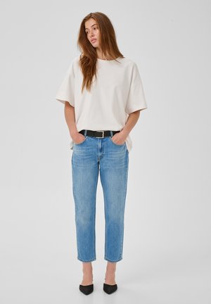Jonge vrouw met lang haar staat met handen in haar zakken, draagt een wit oversized T-shirt, blauwe spijkerbroek met zwarte riem en zwarte puntige schoenen.
