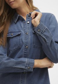Jeansskjorta i medelblå färg med krage, två bröstfickor med lock och metallknappar. Långa ärmar med knäppning vid ärmsluten.