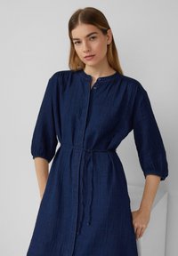 Vestito button-up di un blu navy scuro, con colletto alla mandarino, maniche a tre quarti e vita legata. Tessuto morbido e testurizzato con una vestibilità rilassata.