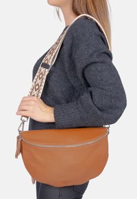 Sac bandoulière en cuir marron, de forme arrondie, avec une surface texturée et une bandoulière décorative à motif. Détails de fermeture éclair visibles.