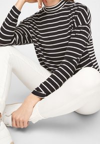 Musta-valkoisi raidoitettu pitkähihainen paita, jossa on korkea kaulus, yhdistetty valkoisiin tiukkoihin farkkuihin ja valkoisiin lenkkareihin, esittäen rentoa tyyliä.