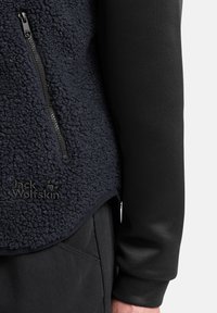 Fleecejacke in Dunkelblau, mit strukturiertem Oberflächen, schwarzem Reißverschluss und Logo; kombiniert mit einer glatten schwarzen Langarmschicht darunter.