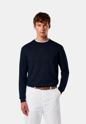 Giovane uomo con capelli ricci indossa un maglione blu scuro, pantaloni bianchi e cintura marrone, in piedi davanti a uno sfondo chiaro e uniforme.