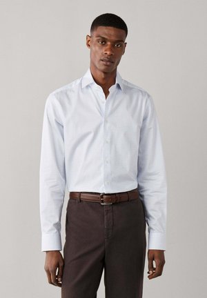 Man wearing a light blue checkered button-up shirt tucked into dark brown pants with a brown leather belt, standing against a plain background.