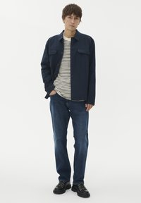 Mornarska jakna z gumbom in dvema žepoma na prsih, oblečena čez belo in mornarsko prugasto srajco. Slim-fit modri kavbojki in črne vezalne čevlje.