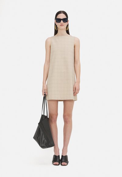 Beige kariertes Mini-Kleid mit quadratischer Form und ärmellosem Design. Accessoires sind schwarze übergroße Sonnenbrillen, goldene Creolen und eine schwarze Tragetasche.