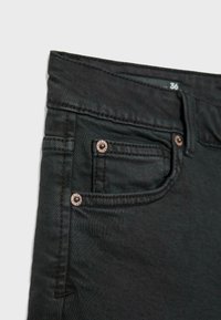 Jean en denim noir en gros plan montrant la poche avant, les rivets, la boucle de ceinture et l’étiquette de taille marquée 36 à l’intérieur de la ceinture.