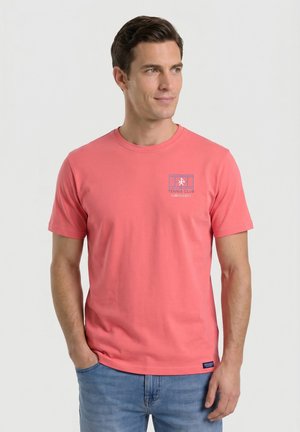 Hombre que lleva una camiseta de manga corta color coral rosa con un pequeño logo de club de tenis en el pecho izquierdo y vaqueros azul claro, de pie con una mano en el bolsillo.