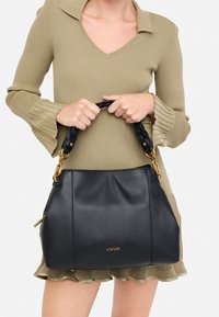 Sac à main en cuir noir avec une surface texturée, une anse tressée et des accens en ton doré. Le sac a une forme douce et structurée.