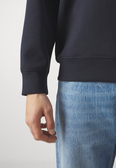 Felpa blu navy con polsini a costine, abbinata a jeans in denim azzurro. Il tessuto appare liscio; le cuciture sono visibili lungo i bordi.