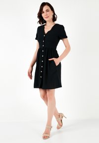 Abito nero a maniche corte con scollatura a V, chiusura con bottone anteriore, gonna svasata e tasche laterali. Tessuto liscio, lunghezza al ginocchio, abbinato a tacchi beige.