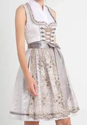 Woman wearing a sleeveless traditional dress with lace-up bodice, white blouse, and a floral embroidered apron tied with a wide satin ribbon.