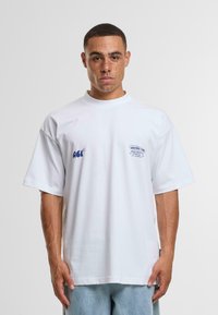 Camiseta blanca de algodón con cuello alto, corte relajado y mangas cortas. Presenta un logo y texto en azul en el frente. Textura suave.