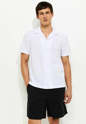 Młody mężczyzna w białej, krótko rękawowej koszuli zapinanej na guziki oraz czarnych szortach do kolan, stoi z jedną ręką w kieszeni.