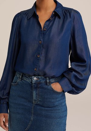 Kvinde iført en mørkeblå skjorte med knapper og puffede ærmer, stukket ned i en højtaljet blå denimnederdel. Den ene hånd i lommen.