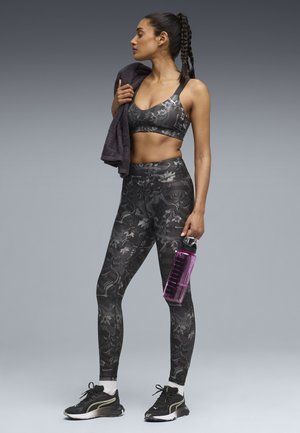 Sort sports-bh og leggings med mønster lavet af strækstof, kombineret med sorte træningssko. Holder en klar pink vandflaske og et gråt håndklæde.