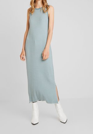 Femme portant une robe maxi sans manches rayée bleu clair et gris avec une fente sur le côté, associée à des bottines blanches, debout sur un fond uni.