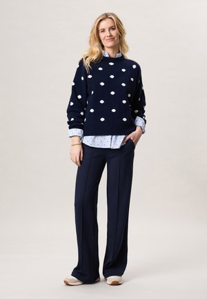 Vrouw staand, gekleed in een marineblauwe trui met witte stippen over een gestreept shirt, marineblauwe wijde broek en beige sneakers, glimlachend met één hand in de zak.