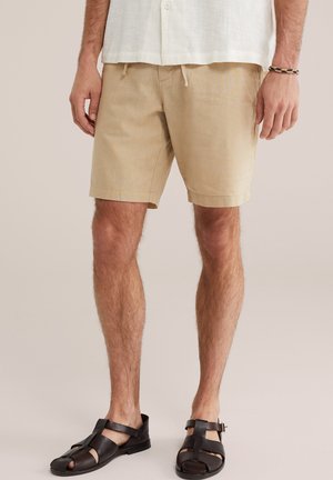 Meesmodell kannab beežikaunistustega lühikesi pükse, valget särki, mustad nahast sandaale ja punutud randmerõngast, seisab ühevärvilise tausta ees.