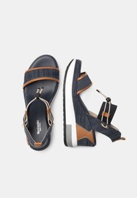Sportsandalen mit Obermaterial aus Denim, braunen Lederelementen, verstellbaren Riemen und einer weißen Gummisohle. Merkmale sind eine strukturierte Oberfläche und ein flaches Design.