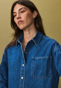 Blauwe denim shirt met een knoopsluiting aan de voorkant, puntcollar, één borstzak en subtiele textuur.