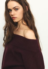Bordový pletený sveter s odkrytými ramenami, so žebrovaným výstrihom a jemnou textúrou, ktorý sa jemne zhadzuje cez jedno rameno.