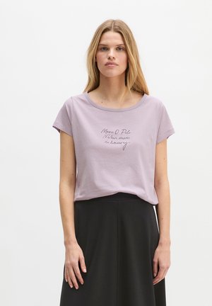 Jeune femme aux longs cheveux blonds portant un t-shirt violet clair avec un texte et une jupe noire, debout devant un fond blanc uni.